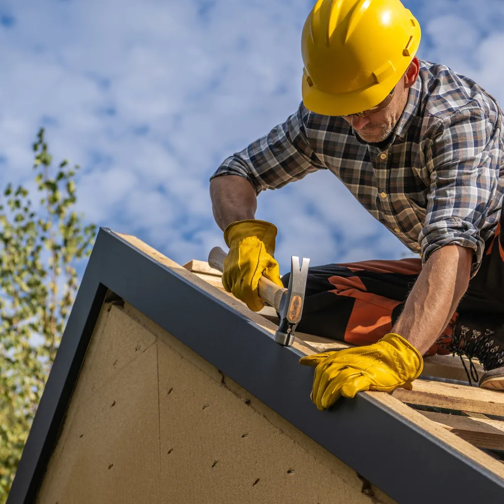 Roofer in safety gear hammering roof framing under a blue sky